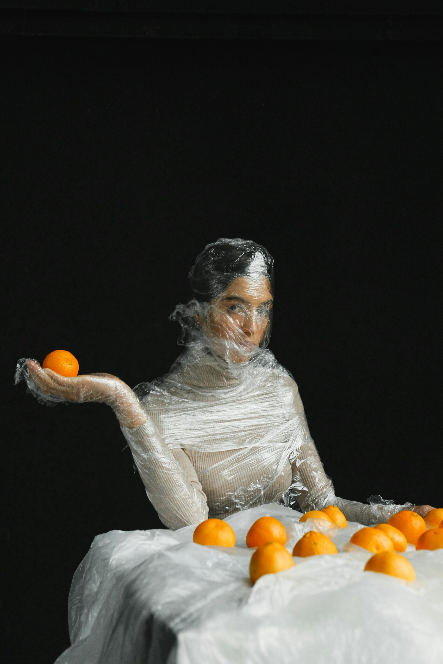 A woman covered in plastic, holding an orange, highlighting pollution and waste.